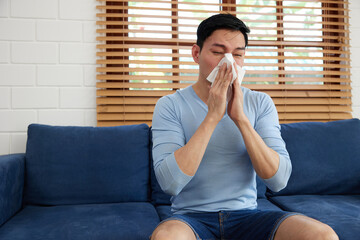young man having a cold, using tissue paper and blowing his nose on sofa