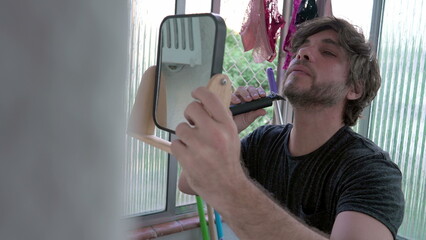 Multi-tasking young man grooming beard with trimmer while holding small mirror at his apartment...