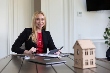 Smiling businesswoman realtor sits at table with papers in modern office. Blonde young lady wears...