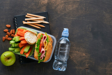 Healthy food for school lunch concept. Healthy bread, meat sandwich with cheese, apple, fresh cucumber, carrots, nuts in container, water and yogurt on dark background. Back to school concept.