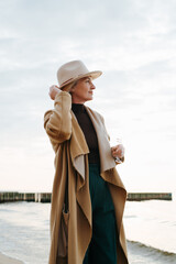 Elegant mature lady in coat touching round brown hat standing on seashore outdoors, senior woman enjoying sea looking at horizon