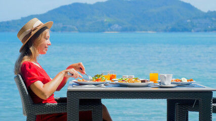 Woman relaxes in luxury hotel, enjoying sea view. Female traveler savors a healthy breakfast in a resort restaurant on a tropical beach, with the turquoise ocean. Thailand vacation.