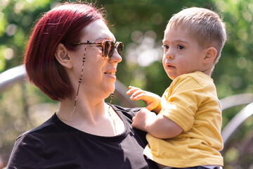 Happy mother in sunglasses holding cute little son with Down syndrome in arms redhead woman with...