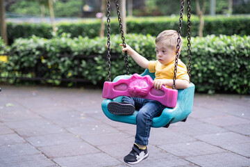 Little blond boy with Down syndrome enjoys riding on safe children swings against lush bushes cute...