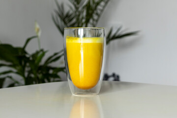 Golden milk in a transparent cup against the background of room plants and a white room