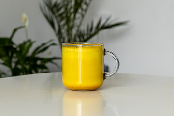 Golden milk in a transparent cup against the background of room plants and a white room