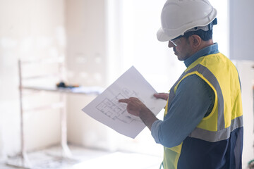 Specialist examining interior project drawing standing against construction safety equipment man in professional uniform stands in room controlling renovation process in apartment