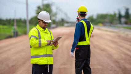 Asian engineer holding tablet and foreman using walkie talkie to control road construction, against backdrop of road under construction