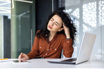 Overtired hispanic business woman working inside modern office, office worker with laptop having severe headache.