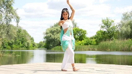 Beautiful Indian woman dancer wearing traditional blue white dress dancing dance on wooden pier by the river.