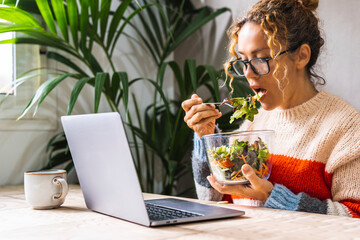 Woman on diet eating fresh salads from transparent bowl while using laptop at home watching online...
