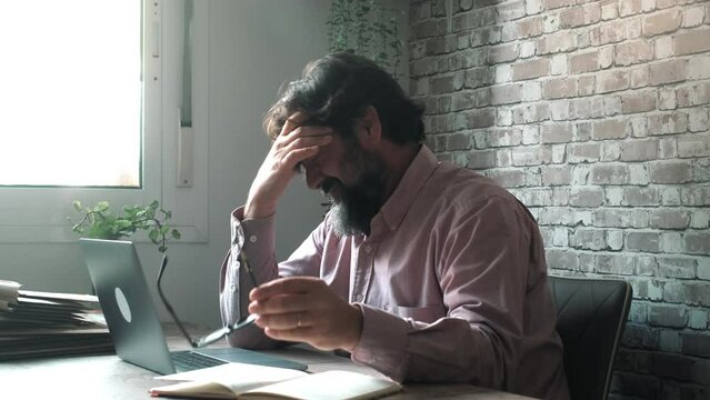 One overworked tired man taking off his glasses and touching his painful head for migraine headache in front of a laptop sitting at the desk. Concept of stress and head ache disturb working computer