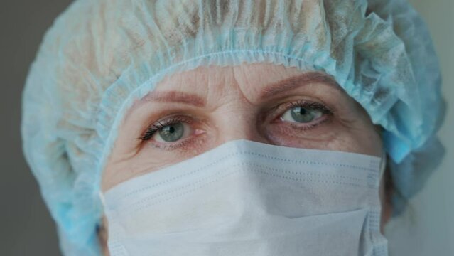 Close-up portrait of an elderly woman doctor medical worker in a mask and cap after surgery. Tired eyes of an adult female doctor. High quality 4k footage