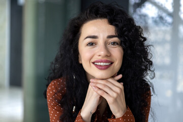 Close-up portrait of Latin American woman, businesswoman boss smiling and looking friendly at camera, female worker with curly hair successful inside office.