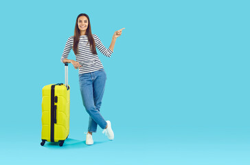 Image of a beautiful brunette with long straight hair in a striped black and white jumper, holding a large yellow suitcase in one hand, and pointing to an empty space for advertising with the other.