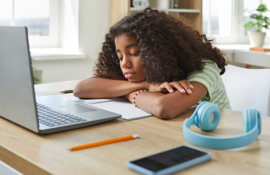 Elementary school child gets bored and tired during online class. Sad diverse Afro American student girl sitting at her desk with laptop and notebook lays head on arms and thinks about something else