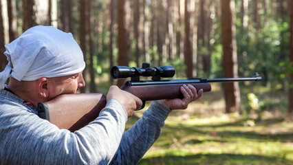 Shooting in the forest from an air rifle with an optical sight. Sunny summer day, back view. - obrazy, fototapety, plakaty