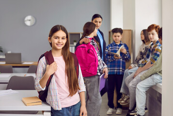 Portrait of happy girl elementary student standing with backpack. Cute schoolgirl wearing casual clothes posing in classroom on background of her classmates. Smiling schoolgirl looking at camera