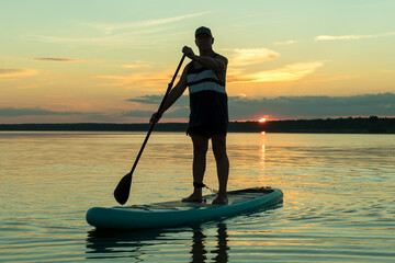 A man in shorts on a SUP board with a paddle at sunset swims in the water of the lake in the glare from the setting sun.