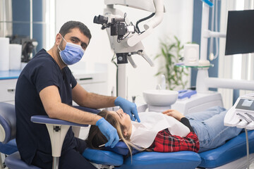 Successful dentist in medical mask sitting near female patient on chair and dental microscope looking in camera professional equipment in modern dentistry clinic