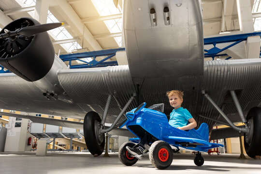 Portrait of cute little blond happy kid boy enjoy have fun play riding pedat toz plane model against vintage airplane background in museum hangar. Future child profession dream concept