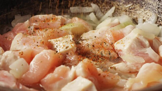 Pieces of chicken fried in a cast-iron skillet in oil with onions sprinkled with spices. Close-up