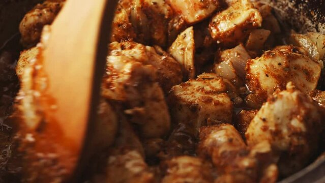 Pieces of chicken fried in a cast-iron skillet in oil with onions and spices are mixed with a wooden spatula. Close-up