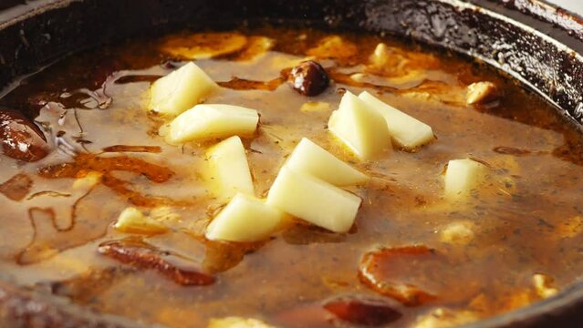 Pieces of chicken are fried in a cast-iron skillet in oil with onions and spices in the broth, where chopped potatoes fall. Close-up