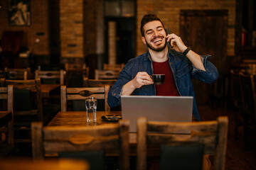 Young businessman owner or manager of a cozy bar and cafeteria, using a phone and a laptop computer to prepare everything for the upcoming day
