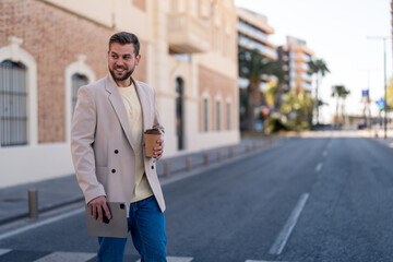 Young stylish modern business man walking with coffee cup and digital tablet looking away and smiling while crossing the street in the city. Photo with copy space.