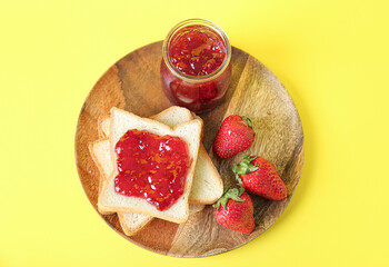 Wooden plate with toasts, jar of tasty jam and fresh strawberry on yellow background