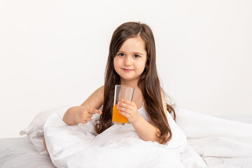 Happy little girl child in pajamas with a glass of fresh orange juice sitting at home in bed. Good morning