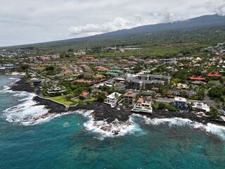 View of coastal Kailua-Kona on the Big Island of Hawai'i. Aerial drone looking along the coast with palm trees, houses, condominiums, and blue and green waters with volcanic rock and coral.