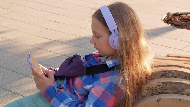 Blonde child headphones checkered shirt listen to music podcast smartphone outside city street urban lifestyle. Concentrated girl online audiobook social media. Mental health application Stress relief