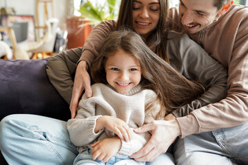 Portrait of happy smiling adorable little daughter child in gentle embrace of young loving parents enjoying family lifestyle games at cozy home living room during spending weekend fun time together