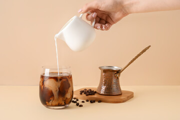 Woman pouring milk from jug into glass with ice coffee on beige background