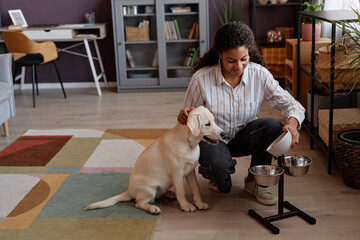 Full length portrait of smiling black woman feeding puppy at home and using stand with metal bowls
