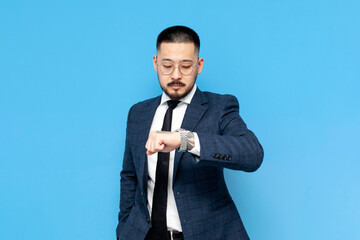 successful asian businessman in suit and glasses is late and looks at his watch on blue isolated background - Powered by Adobe