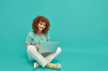 Foto op Canvas Smiling cheerful smart young ginger woman wearing casual teenage clothes using laptop computer sitting on floor in lotus pose looking at camera isolated over turquoise background with copy space © Stock 4 You