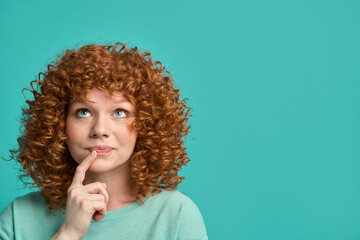 Headshot portrait of thoughtful pensive young ginger woman with curly hair holding finger on lips...