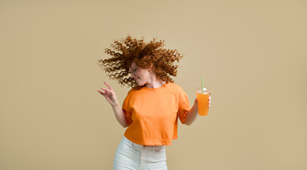 Happy carefree young wavy-haired ginger woman dancing holding fresh juice drink cocktail in hand...