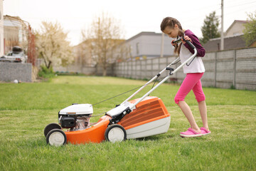 Cute teenage child girl working in garden, mowing green grass with lawn-mower on backyard on summer holiday. Concept of pocket money, first salary. Special equipment for landscape, terrain. Gardening