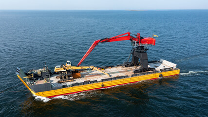 Yellow barge loaded with tracked harbour material handling cranes, being pulled across the open ocean. Aerial side view.