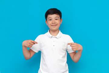 little asian boy in white t-shirt points to himself on blue background, korean child advertises copy space