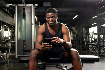 young athletic african american man sits in dark gym and uses smartphone, sports guy types online on the phone