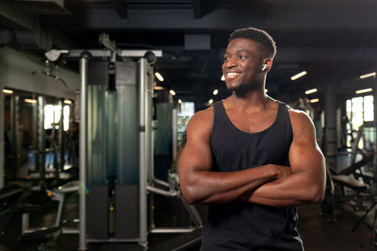 young African American man stands with crossed arms in dark gym and smiles, fitness trainer stands in fitness club