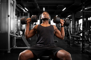 athletic african american man trains in dark gym, athletic guy lifts heavy dumbbells in fitness club