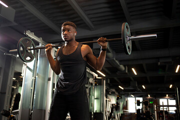 Fototapeta na wymiar african american man lifts barbell in dark gym, sports guy trains in fitness room and goes in for sports