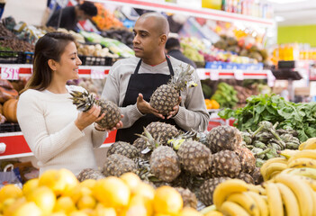 Latin woman choosing ripe pineapples in supermarket and shop worker helping