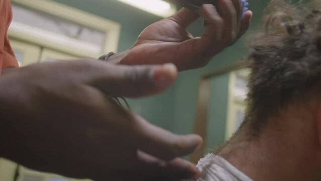 Close-up shot of hands of black barber trimming neckline of male client with hair clippers and comb while working in barbershop